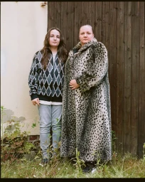 Two people posing outdoors in front of a rustic wooden door and pale wall with tall grass at their feet.