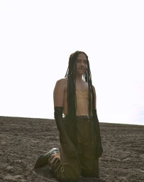 Person with long braided hair kneeling in a plowed field wearing a beige top and dark pants