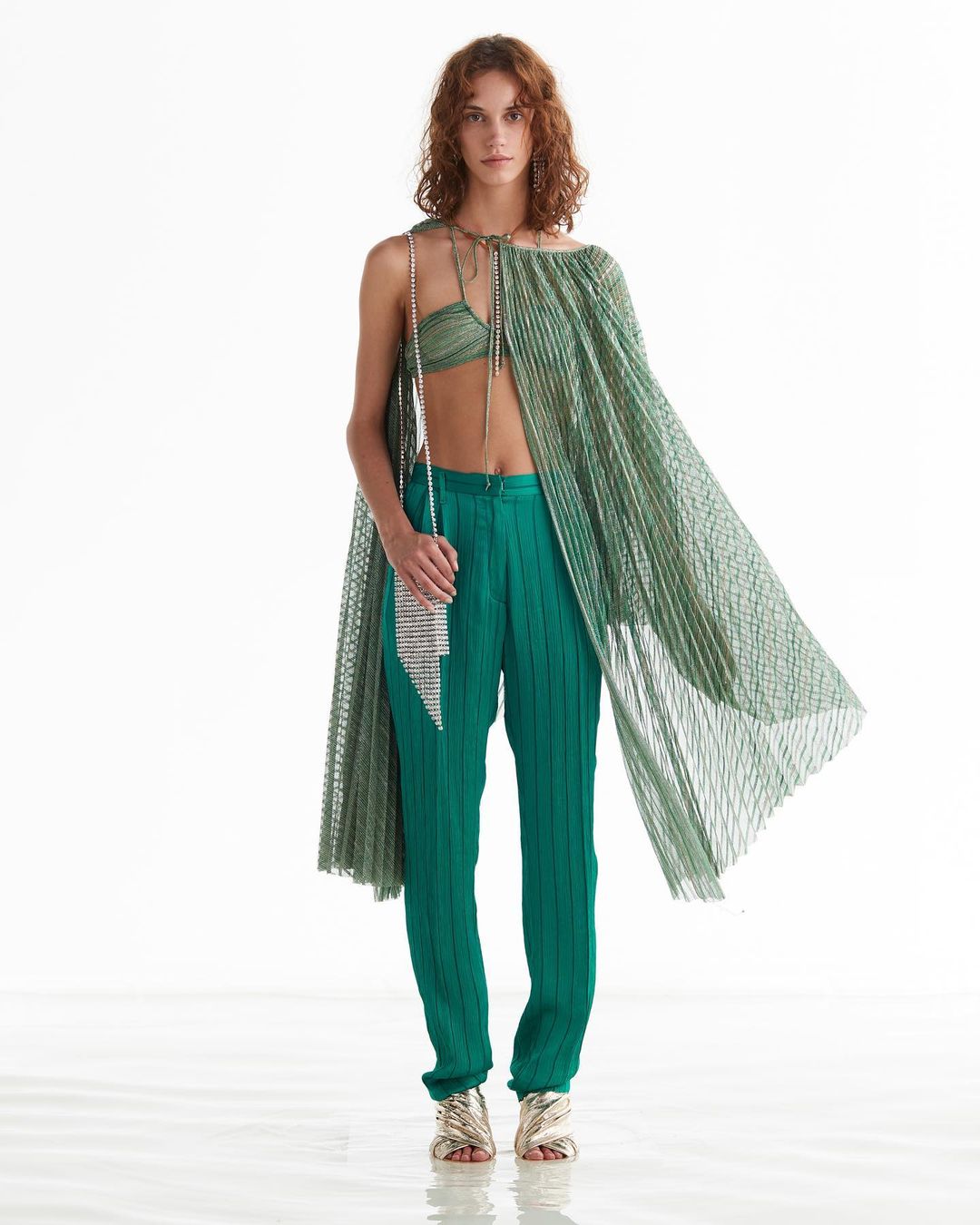 Fashion shot of a woman in pleated green top and pants with a silver purse, posing in a studio with a white backdrop.