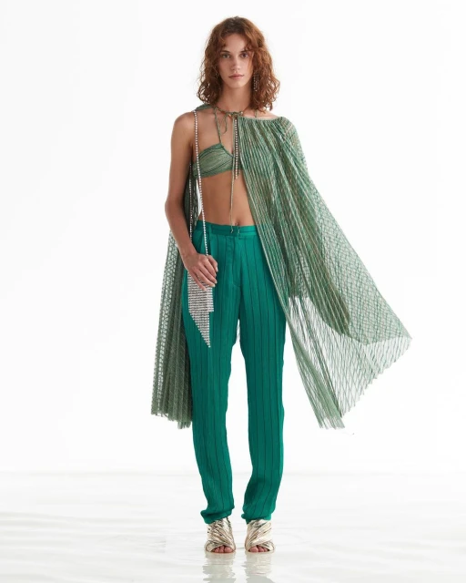 Fashion shot of a woman in pleated green top and pants with a silver purse, posing in a studio with a white backdrop.