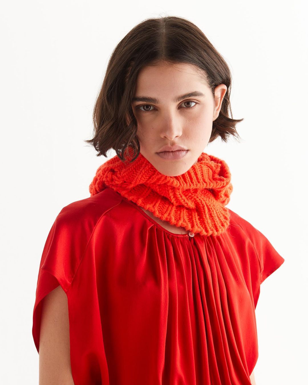 A woman with short dark hair is posing indoors, wearing a red shirt and an orange knitted neck warmer against a white background.
