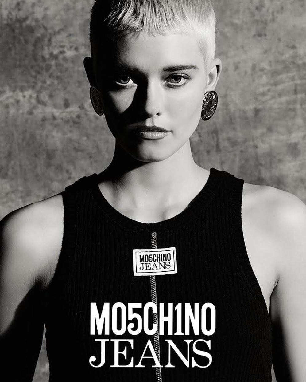 Close-up portrait of a white woman with short blonde hair and dark earrings wearing a black ribbed tank top against a textured concrete background