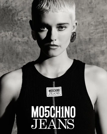Close-up portrait of a white woman with short blonde hair and dark earrings wearing a black ribbed tank top against a textured concrete background