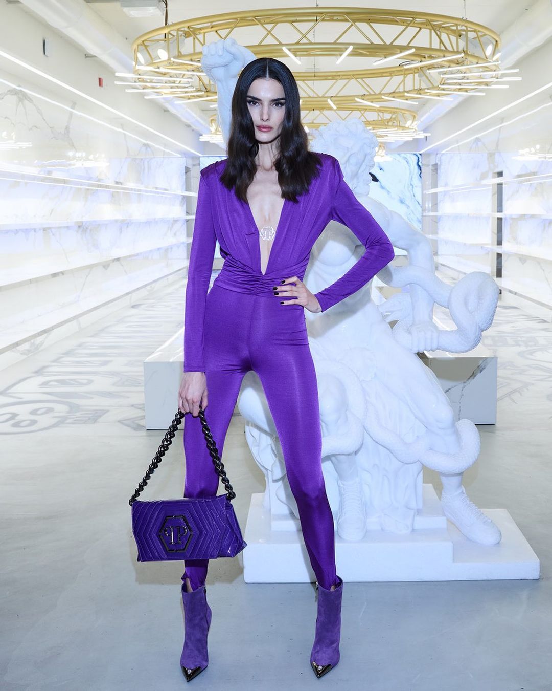 Fashion model poses in a purple jumpsuit with a matching handbag and boots in a brightly lit retail setting with decorative sculptures.