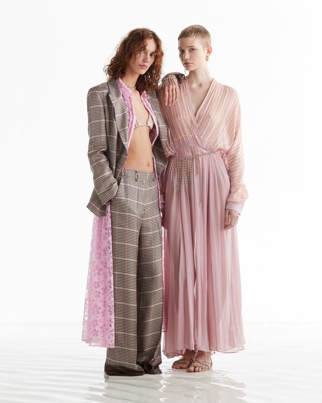 Two female models posing together against a plain white studio background wearing patterned plaid suit and pink pleated dress.