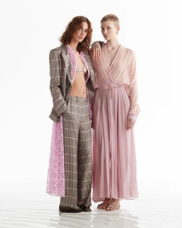 Two female models posing together against a plain white studio background wearing patterned plaid suit and pink pleated dress.