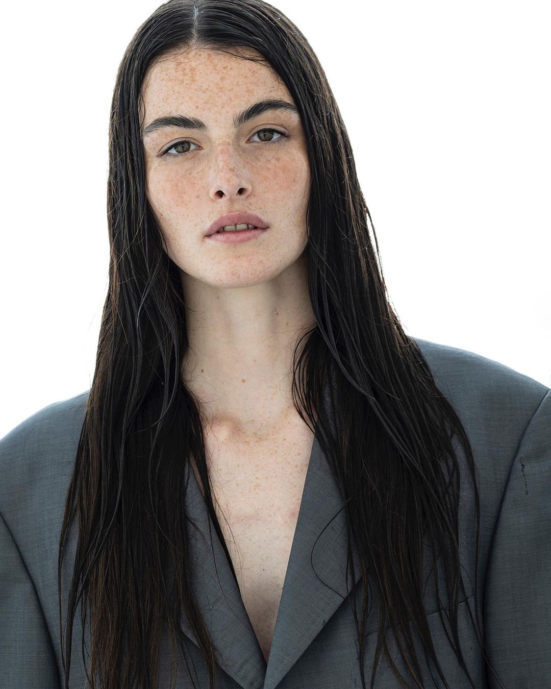 Close-up portrait of an adult female model with long, dark, wet hair, light skin, and freckles, wearing a grey blazer against a white background.