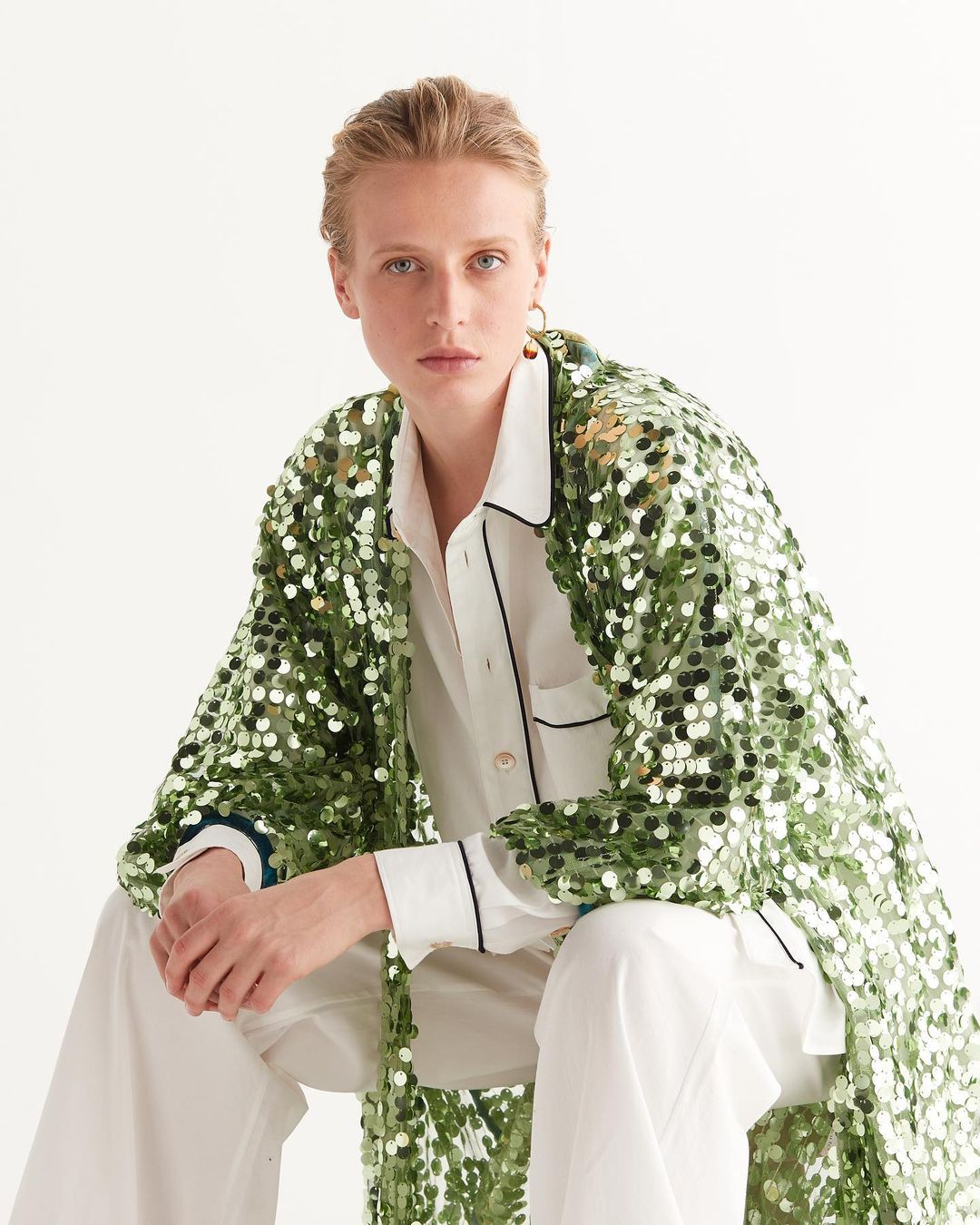 Woman wearing a sequin robe and white pajamas sitting in a photo studio. The lighting is bright and the image is fashion-focused.