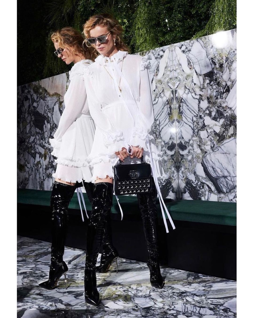 Fashion model wearing a white mini dress and black thigh-high boots poses in front of a marble wall with greenery in the background.