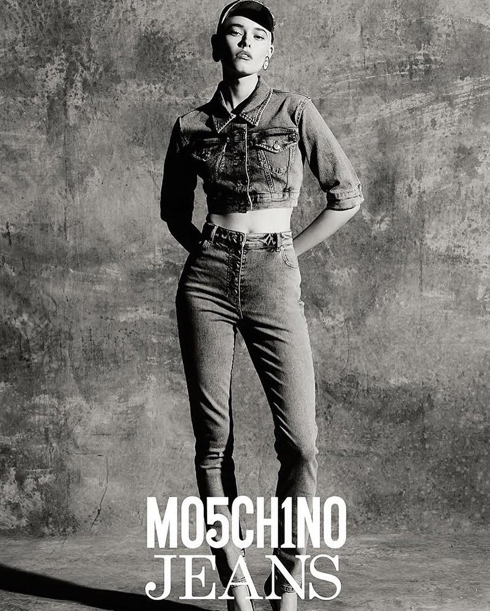 A slender fashion model poses in a grey-scale studio shot, wearing a denim jacket and jeans by Moschino, hands behind back.