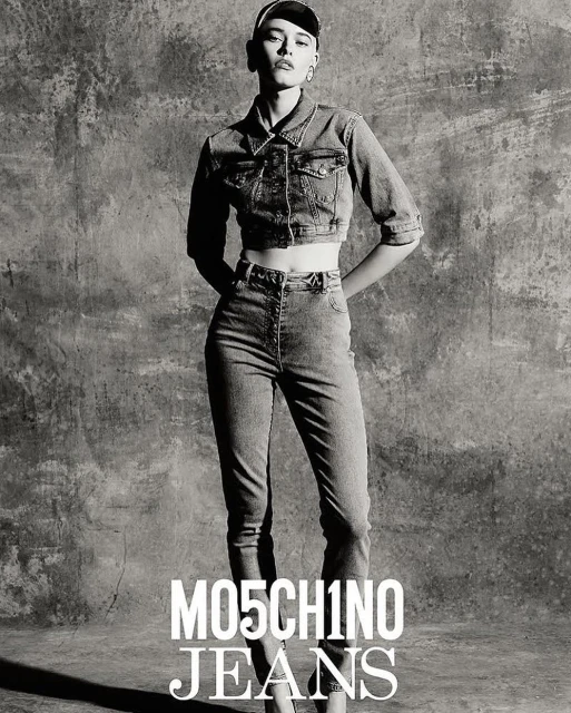 A slender fashion model poses in a grey-scale studio shot, wearing a denim jacket and jeans by Moschino, hands behind back.