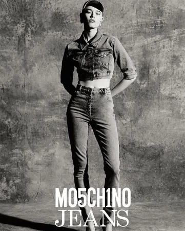 A slender fashion model poses in a grey-scale studio shot, wearing a denim jacket and jeans by Moschino, hands behind back.