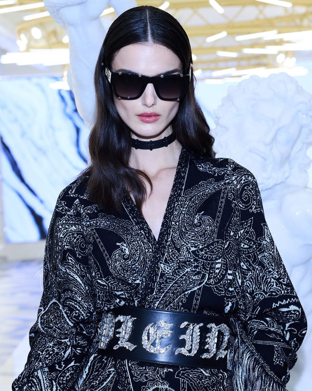 A woman with long dark hair wearing sunglasses, a paisley print jacket, and a thick belt stands indoors in front of a white marble sculpture.