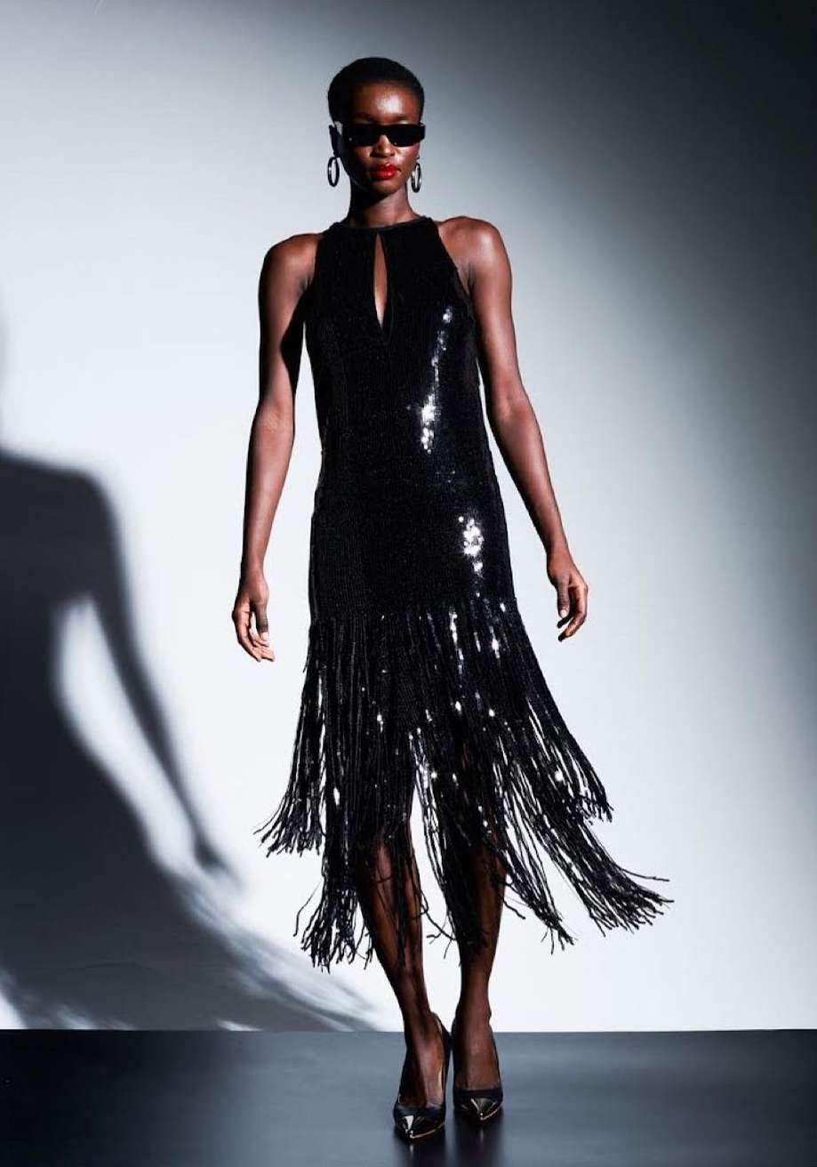 A Black woman poses in a shimmering, knee-length black fringe dress with sunglasses, standing in a light grey studio environment.