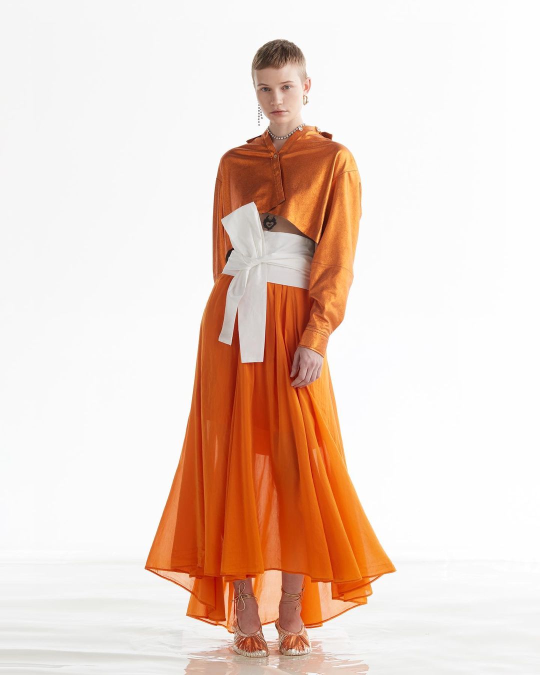 A fashionable woman poses in an orange two-piece outfit with a white obi belt, showcasing the elegant design against a bright backdrop.