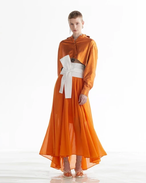 A fashionable woman poses in an orange two-piece outfit with a white obi belt, showcasing the elegant design against a bright backdrop.