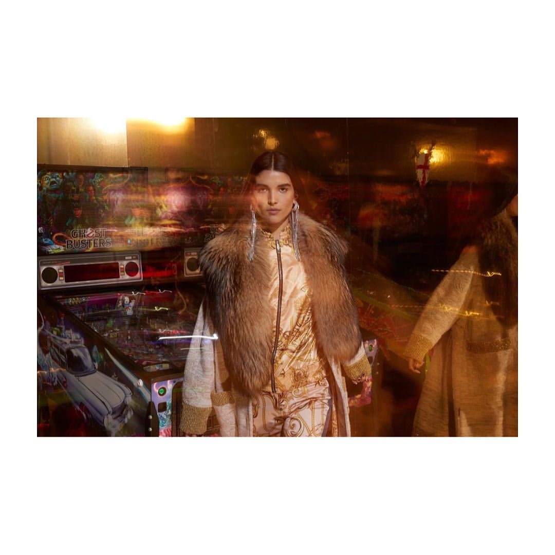 A woman wearing a fur collar coat poses near a Ghostbusters pinball machine with warm lighting and a medium shot in an indoor setting.