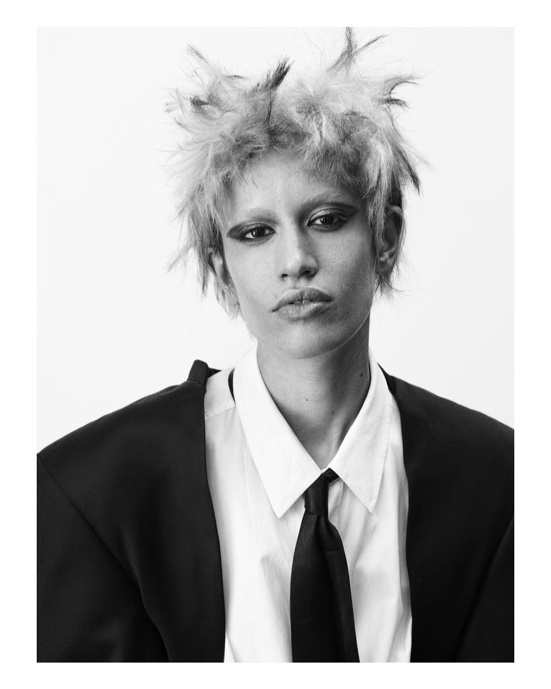 Black and white portrait of a stylish adult with spiky hair, wearing a jacket, white shirt, and tie against a plain background.
