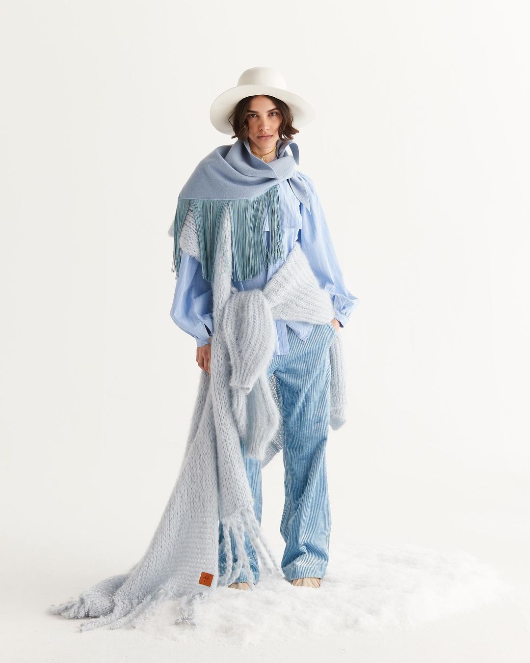 A woman poses in a studio wearing a hat, light blue shirt, wide-leg jeans, and multiple large knit scarves in light blue and gray.
