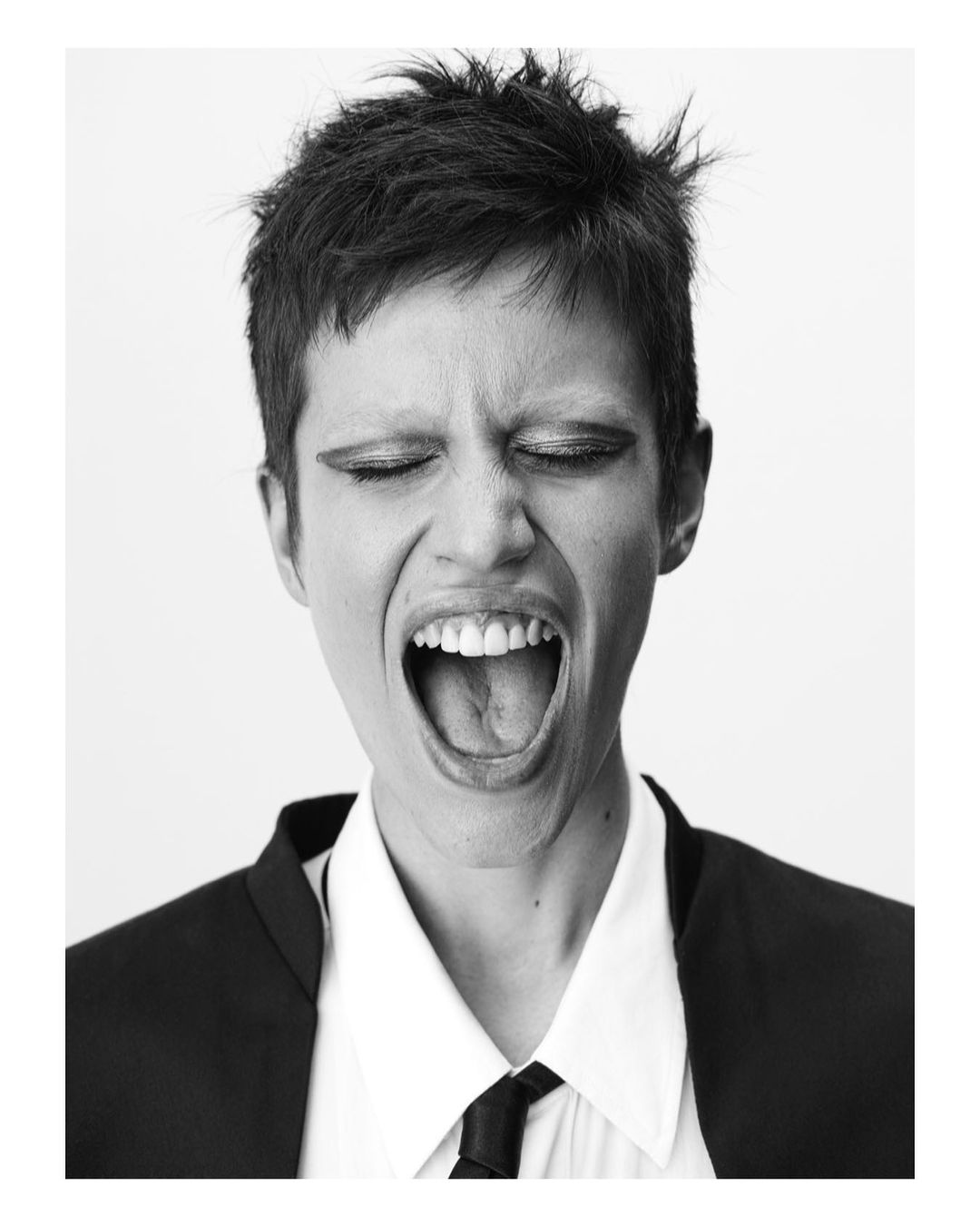 Monochrome portrait of a screaming adult female with her eyes closed, wearing a suit in front of a plain white backdrop.