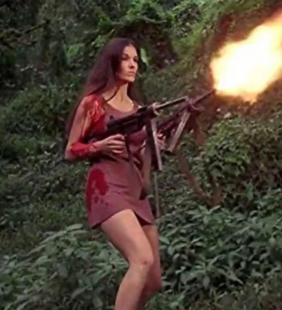 Brunette woman in a short brown dress and what appears to be blood stains fires a machine gun in a lush forest setting.