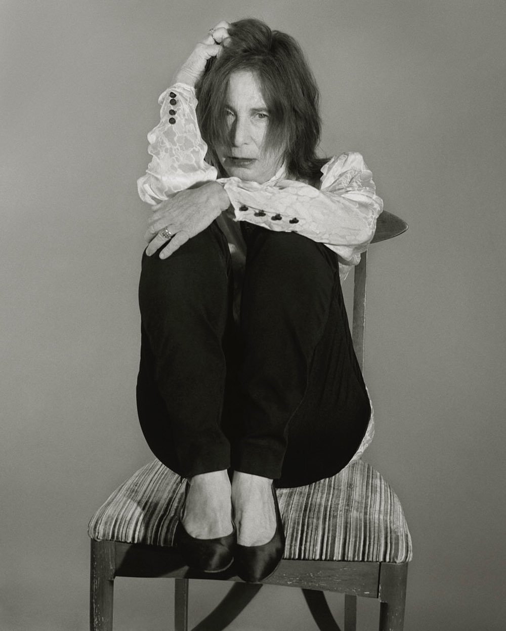 Portrait of mature woman with red hair sitting in chair and hugging her knees in a photo studio, wearing dark pants and a lace blouse.