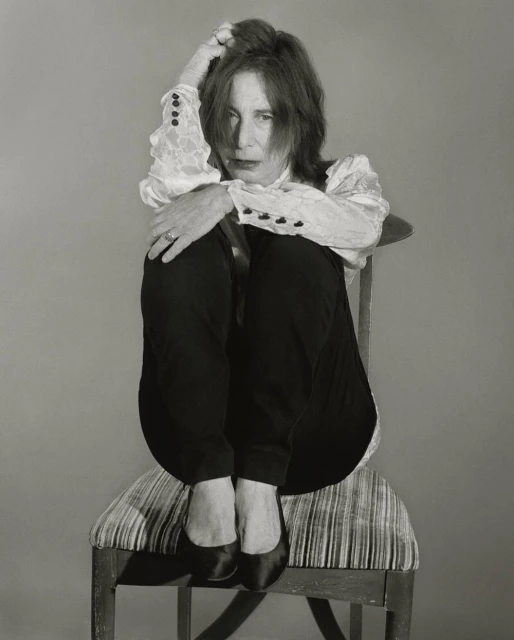 Portrait of mature woman with red hair sitting in chair and hugging her knees in a photo studio, wearing dark pants and a lace blouse.