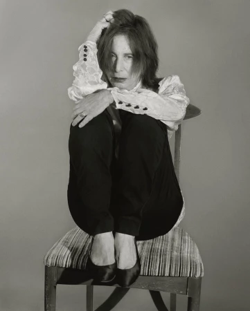 Portrait of mature woman with red hair sitting in chair and hugging her knees in a photo studio, wearing dark pants and a lace blouse.