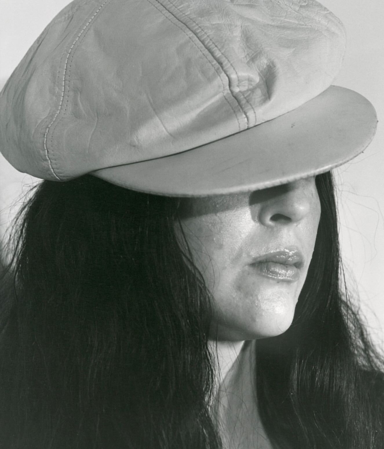 Monochrome portrait of an adult woman wearing a pale newsboy cap, with long dark hair against a neutral grey studio backdrop.