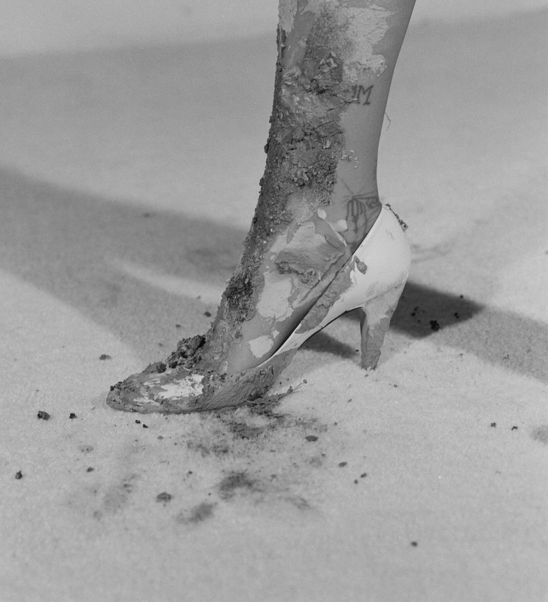 Mud-covered high-heeled shoe on woman's foot, studio shot. Cream-colored heel, ankle tattoo, gray background, monochrome, dramatic lighting.