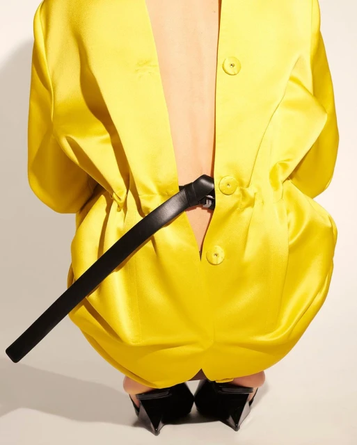 A person wears a yellow oversized blazer, cinched with a black belt, and pointed black shoes, squatting in a studio with a light background.