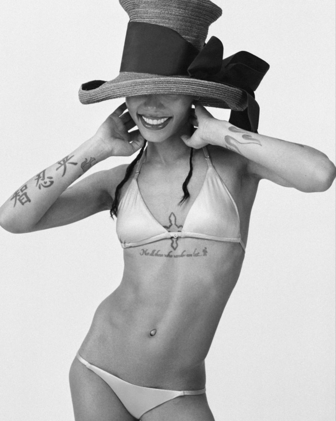Black and white studio shot of a smiling adult woman in a bikini and large hat, posing with hands near her head. Tattoos are visible.