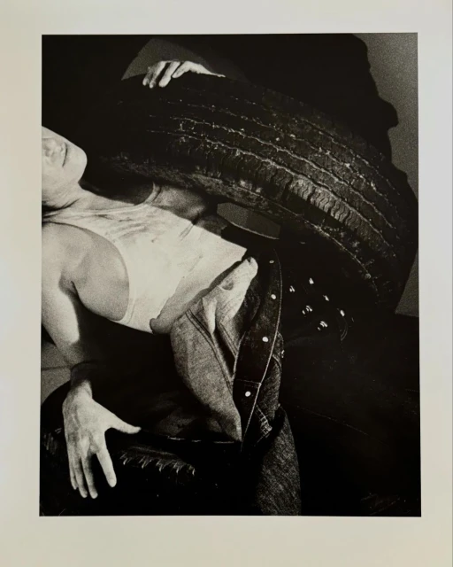 Monochrome photograph of a man holding a large tire, wearing jeans and a light-colored tank top in front of a dark background.