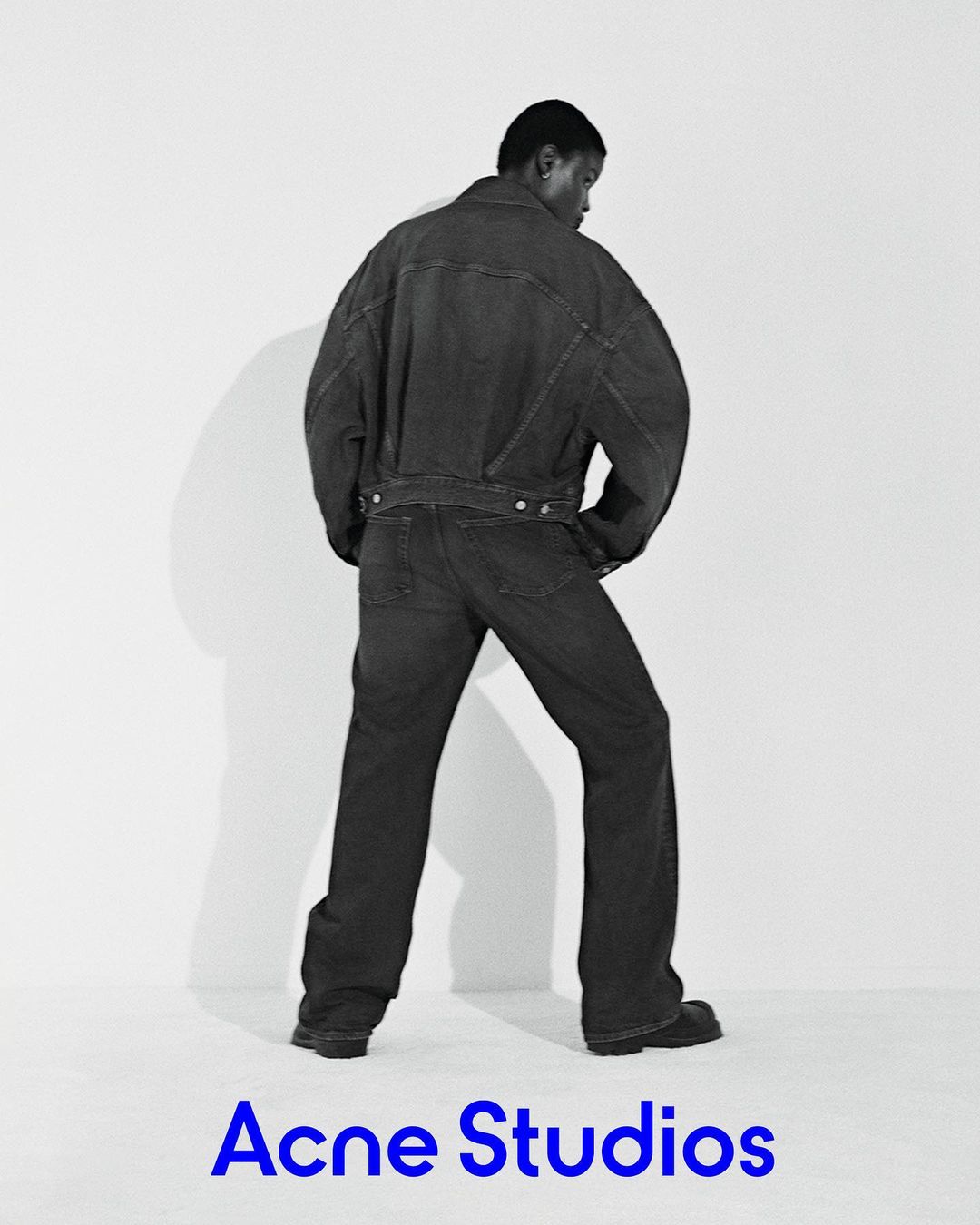 A black male fashion model poses in a dark denim jacket and pants against a white backdrop, presenting a minimalist studio fashion shot.