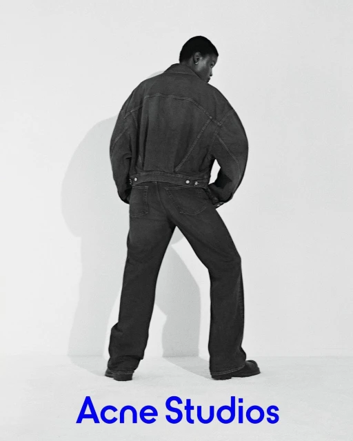 A black male fashion model poses in a dark denim jacket and pants against a white backdrop, presenting a minimalist studio fashion shot.