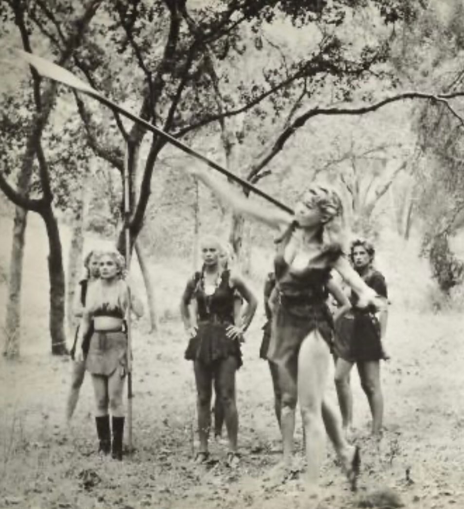 Monochrome image of a woman throwing a spear, with a group of people in primitive attire in a forest setting, in a black and white photograph.