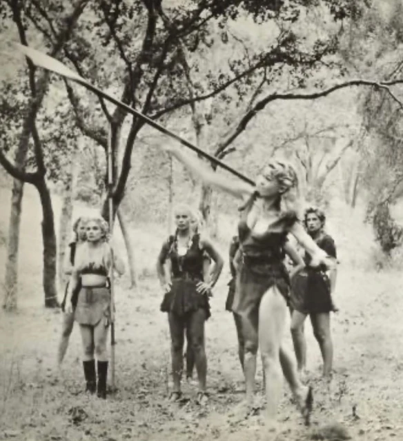 Monochrome image of a woman throwing a spear, with a group of people in primitive attire in a forest setting, in a black and white photograph.