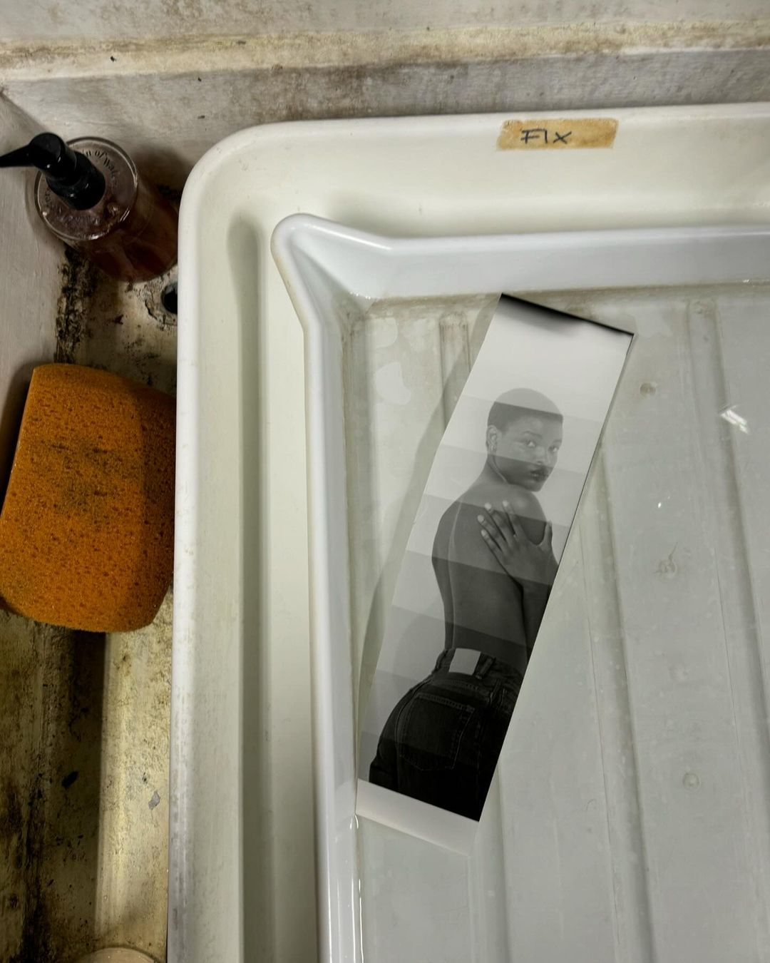 Black and white photograph of a woman in darkroom chemicals, processing film. Development process in a white tray with sponge and soap.