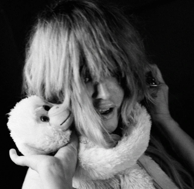 Black and white portrait of an adult with light hair holding a plush monkey, looking at the camera in a dark indoor environment.