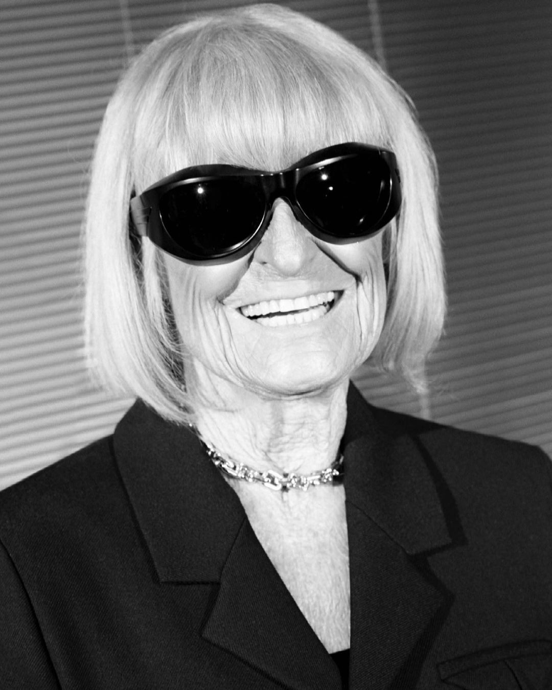 Black and white portrait of an older woman with blonde hair, sunglasses, a blazer, and a necklace smiling into the camera.