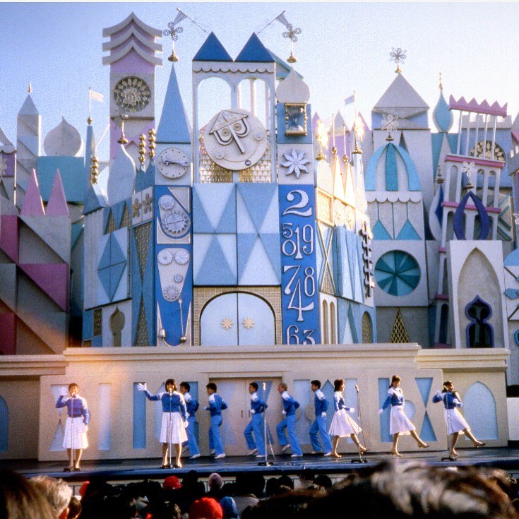 A group of dancers in blue and white costumes perform on a stage in front of a large, colorful, fairytale-like building with towers and clocks.