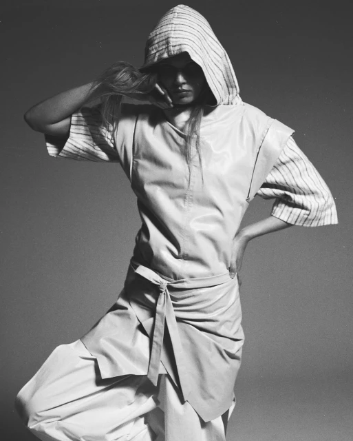 Fashion shot of a woman wearing light-colored streetwear with a hooded top and loose pants, posing in a photo studio with a grey background.