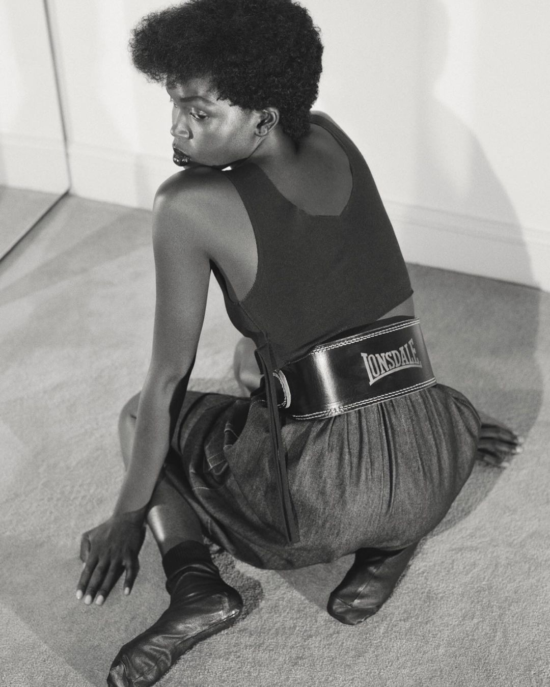 Black and white photo of a slender Black woman in a top and skirt with a Lonsdale belt, seated on the floor, looking over her shoulder.