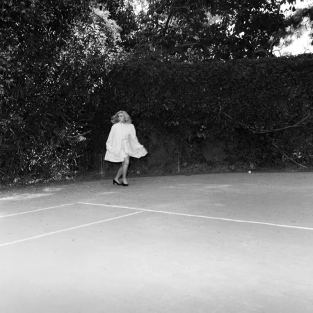 Woman in a white dress and heels dancing on a tennis court with trees and foliage in the background. Black and white, medium shot.