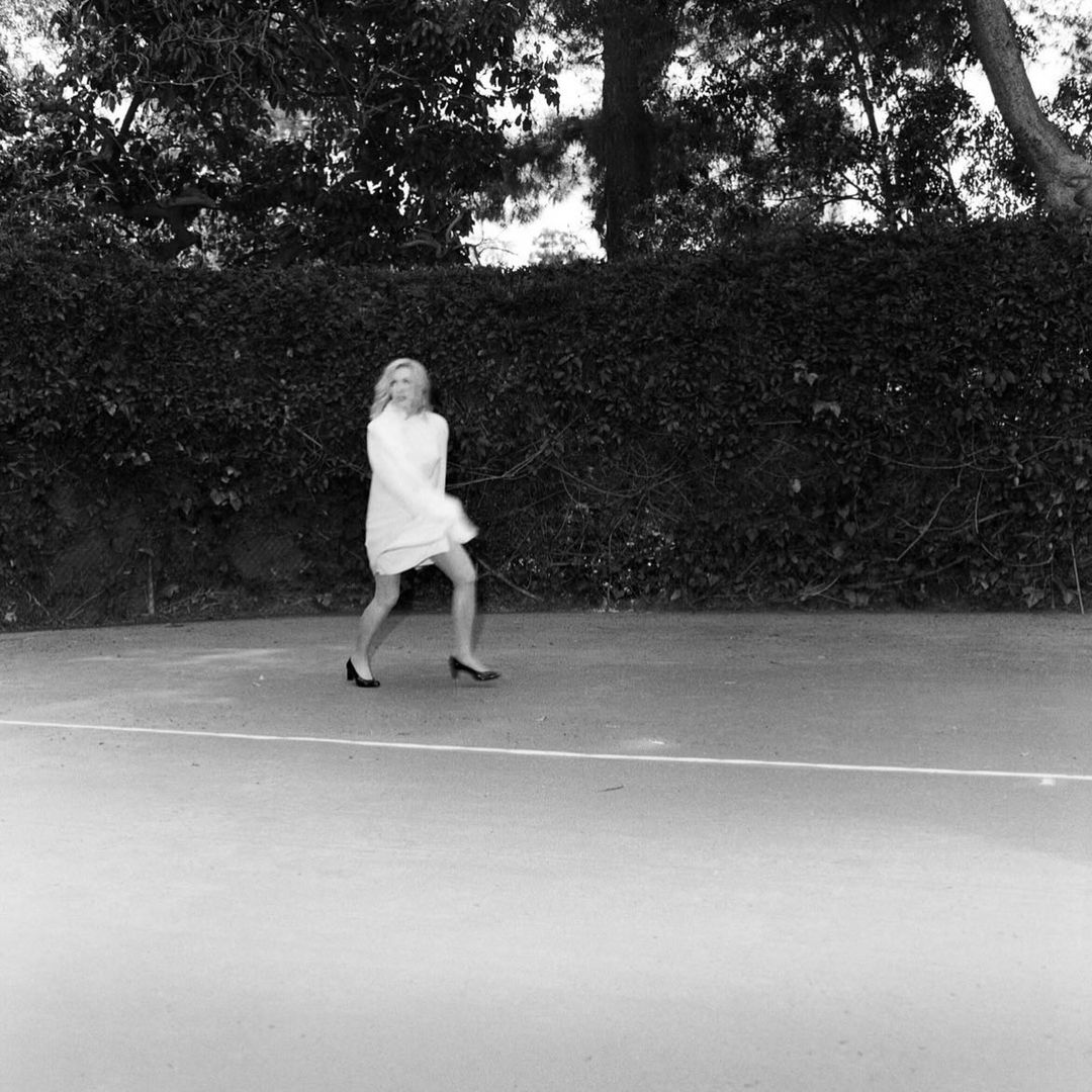 Monochrome shot of an adult female in a white shirt dress and heels, walking across a tennis court with trees in the background.
