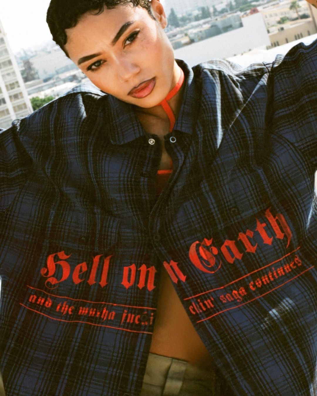 Portrait of a woman wearing a blue plaid shirt with red text on a rooftop with a city skyline in the background on a sunny day.