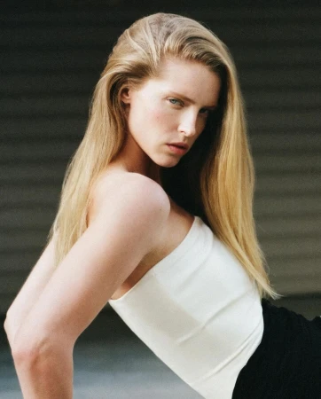Portrait of a blonde woman wearing a white strapless top with long hair looking over her shoulder against a dark corrugated metal background