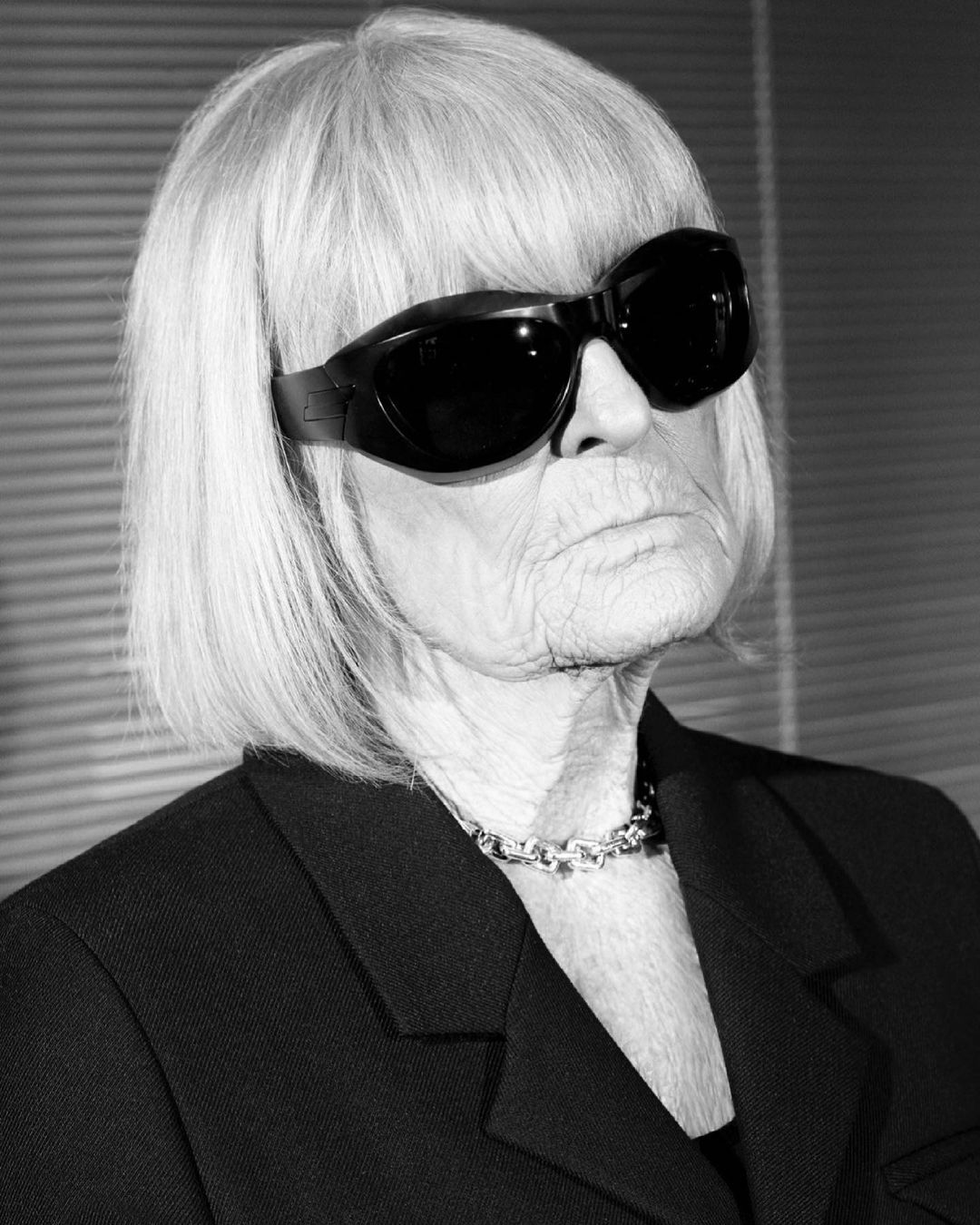 Black and white portrait of an elderly woman with short white hair, wearing dark sunglasses, a black blazer, and a silver necklace.