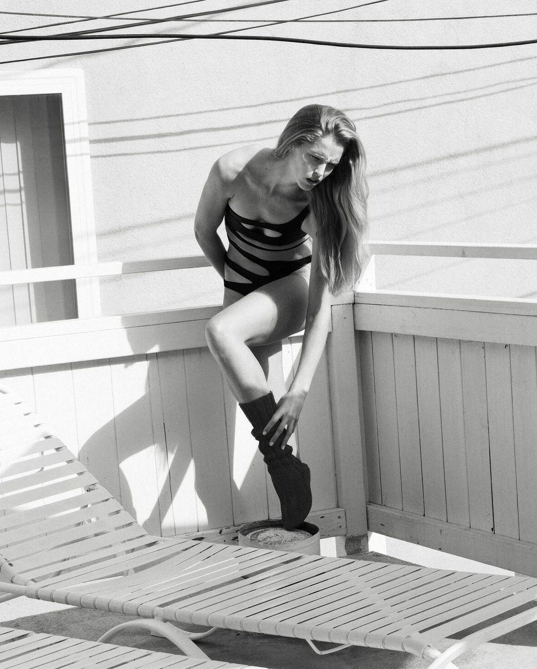 Monochrome portrait of a woman in a black swimsuit and leg warmer leaning against a white railing near a deck chair.