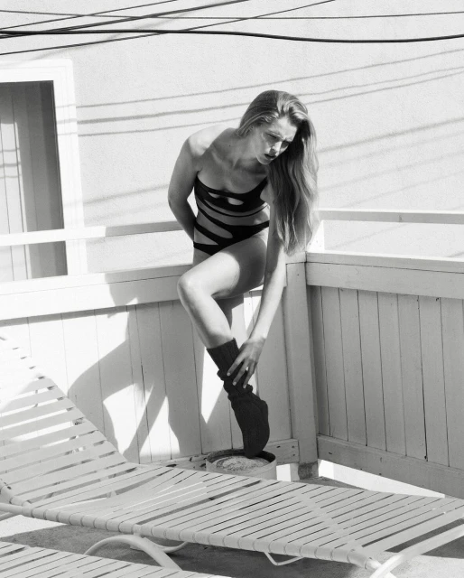 Monochrome portrait of a woman in a black swimsuit and leg warmer leaning against a white railing near a deck chair.